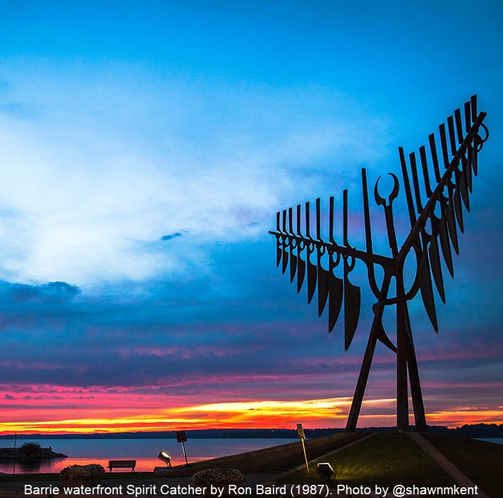 Photo of the Barrie waterfront Spirit Catcher created by Ron Baird in 1987. Photo taken by Canadian photographer Shawn Kent. Photo shows a large metal sculpture of bird-like form against a brightly coloured sunset over Kempenfelt Bay. 