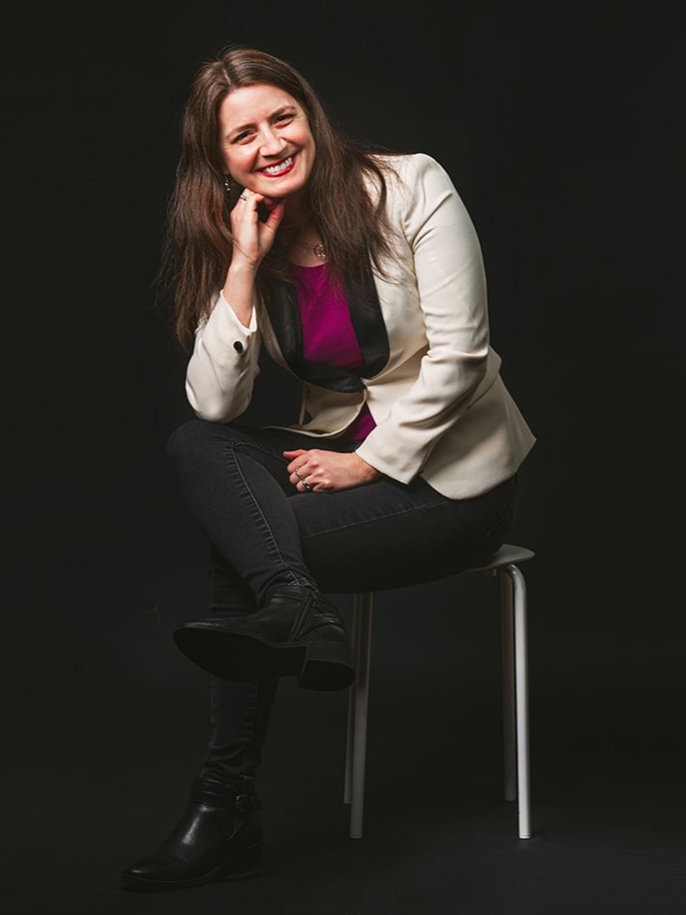 Lorena Aline sitting and smiling against a black background wearing a white and black blazer and magenta shirt.