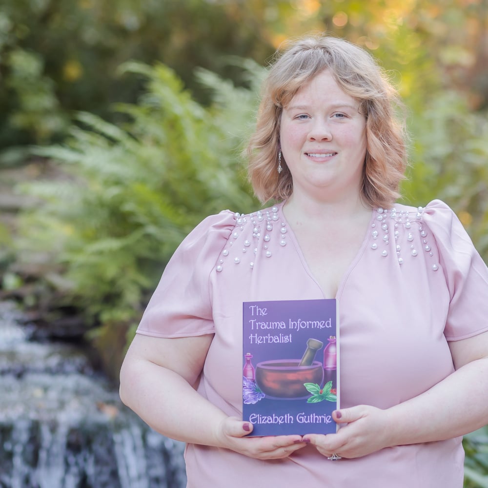 Dr. Elizabeth Guthrie holding her book The Trauma Informed Herbalist in the Birmingham Botanical Gardens