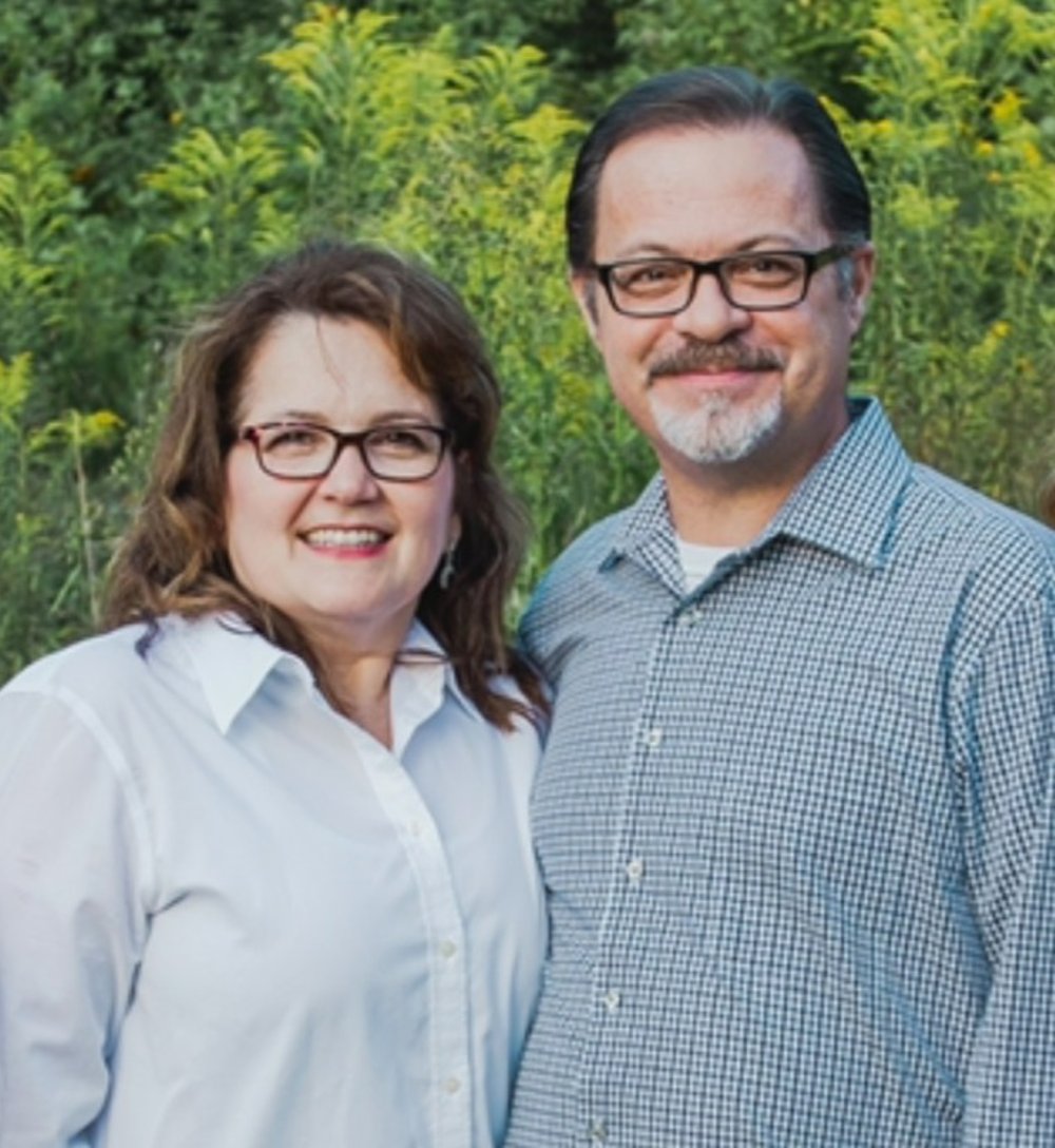 Pastor in a subtle blue plaid shirt with open collar with his wife who is wearing a white button-front blouse
