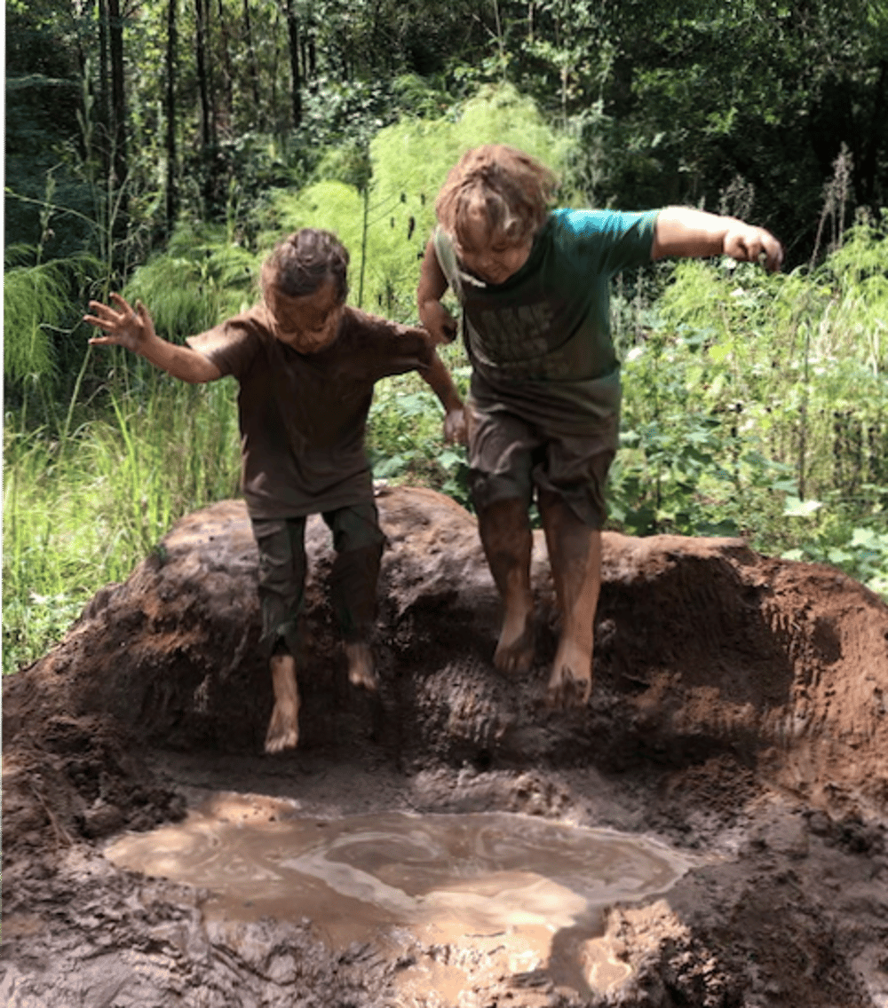 two kids in shirts and shorts jumping from a pile of dirt into a muddy puddle playing the forest  way at one of our nature enrichment programs