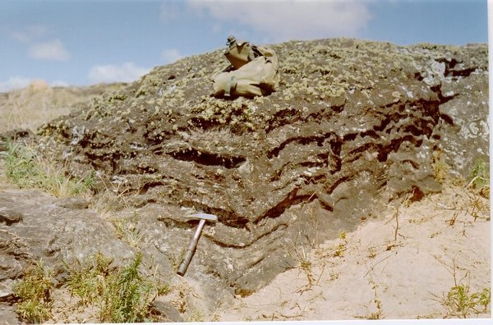 Afloramento de rocha xistosa, com foliação em padrão caixa de ovos. Uma mochila e um martelo servem como estala na foto.