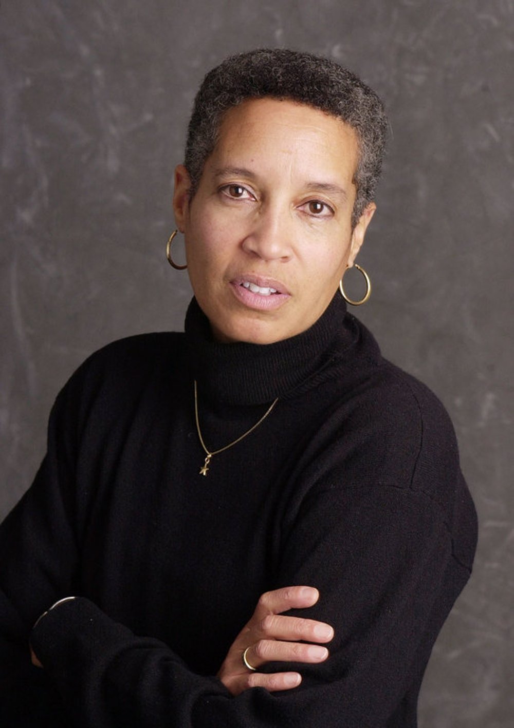 Judge Cordell, staring directly into the camera with her arms folded across her chest. She is wearing gold hoop earrings, a black turtleneck, a gold chain necklace with a small pendant hanging from it
